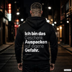 Ich bin das Geschenk. Auspacken auf eigene Gefahr – schwarzer Hoodie mit weißem Backprint, Herren in der Stadt bei Nacht